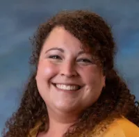 Smiling woman with curly brown hair wearing a yellow top against a soft blue background.