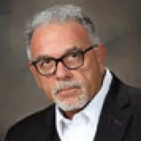 Portrait of an older man with gray hair, glasses, white shirt, and dark blazer against a neutral background.