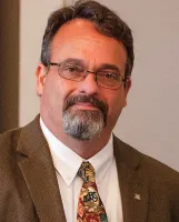 Middle-aged man with glasses, gray beard, wearing a brown suit jacket and colorful patterned tie.