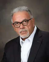 Serious mature man with gray hair and beard wearing black glasses, white shirt, and dark suit jacket against neutral background
