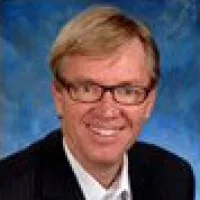 Smiling middle-aged man with glasses and light hair wearing a dark suit and white shirt against a blue background