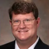 Portrait of a smiling man with glasses, short brown hair, wearing a dark suit and white shirt.