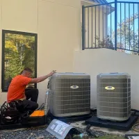 Estes Services Technician in orange shirt installing three Carrier outdoor air conditioning units beside a modern house on sunny day