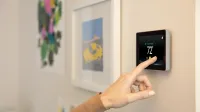 Hand adjusting digital thermostat on beige wall displaying temperature 72 degrees.