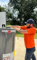 Estes Plumber in orange shirt installing or inspecting a Rheem water heater outdoors on a sunny day.