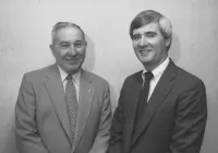 Black and white photo of Napoleon and Tommy Estes in suits smiling and standing against a plain wall background at Estes Heating and Air HQ
