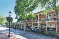 Sunny main street of Roswell, Georgia with historic brick buildings, outdoor seating, green umbrellas, and tree-lined sidewalk with lamp posts.