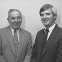 Black and white photo of Napoleon and Tommy Estes in suits smiling and standing against a plain wall background at Estes Heating and Air HQ