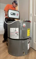 Estes Plumbing Technician in orange installing a gray Rheem water heater inside a home with wooden floor and white doors.