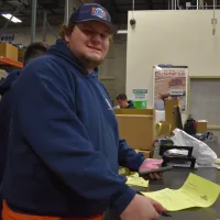 Estes AC Repair Technician in navy hoodie holding documents and phone at busy warehouse counter with coworkers and boxes