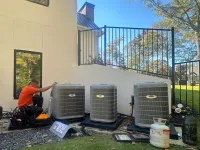 Estes Services Technician in orange shirt installing three Carrier outdoor air conditioning units beside a modern house on sunny day