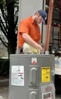 Estes Services Plumber Technician in orange uniform installing or inspecting a Rheem water heater with Energy Guide label in a garage.
