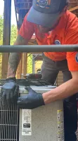 Technician in orange uniform and black gloves servicing outdoor air conditioning unit under wooden porch.