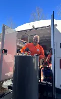 Estes Plumber in orange shirt working on a large water heater tank outside a service van on a clear sunny day.