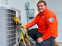 Estes Services Technician in orange uniform kneeling and servicing an outdoor air conditioning unit with gauges and hoses attached.