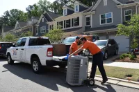 Two Estes technicians unloading a new Carrier air conditioning unit from a white truck in a suburban neighborhood on a sunny day