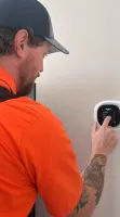 Estes Services Air Conditioner Repair Man in orange uniform and cap adjusts a wall-mounted smart thermostat with a touch screen interface.