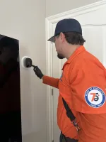 Estes Services Technician in an orange uniform adjusting a smart thermostat set to 72 degrees on a white wall indoors.