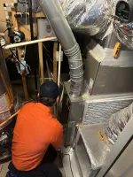 Estes Services Technician in orange shirt inspecting HVAC system components and ductwork in a home's utility room.