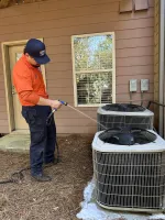 Estes Services Technician in orange uniform cleaning outdoor air conditioning units with a hose near a house with brown siding.