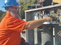 Estes Commercial Technician in blue helmet and orange shirt repairing electrical wiring inside HVAC unit outdoors.
