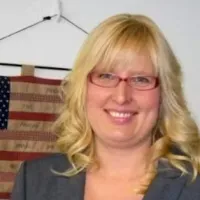 Smiling blonde woman with glasses and curled hair wearing a blazer in front of an American flag.