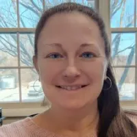 Smiling woman with blue eyes and hoop earrings stands indoors near windows with trees outside.
