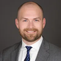 Smiling bald man with beard wearing gray suit and blue polka dot tie against dark background
