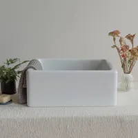 White ceramic farmhouse sink on textured tablecloth with plants and towel in soft natural light.