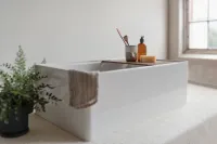 Modern white rectangular bathtub with wooden tray holding soap, toothbrush, and amber bottle beside a potted plant.
