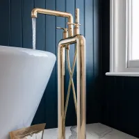 Shiny gold floor-mounted bathtub faucet with running water next to a white tub against dark blue panel wall.