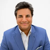 Smiling man with dark hair wearing a blue suit jacket and white striped shirt against a white background