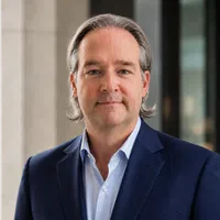 Middle-aged man with gray hair wearing a navy blue suit and light blue shirt in a professional setting.