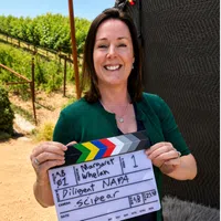 Smiling woman holding a film clapperboard on a sunny vineyard set for a shoot.