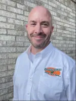 Smiling bald man in white button-up shirt standing in front of gray brick wall with embroidered logo.