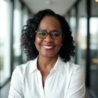 Smiling professional woman with glasses and curly hair wearing a white blouse in a bright office corridor.