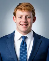 Smiling young man with red hair wearing blue suit, white shirt, and navy blue polka dot tie against gray background