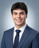 Young man in blue suit, white shirt, and navy tie smiling against a neutral gray gradient background
