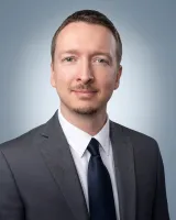 Professional portrait of a man in a gray suit, white shirt, and navy tie against a light blue background.