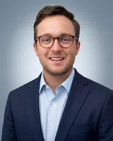 Young man with glasses in a blue suit and light blue shirt smiling against a gray gradient background