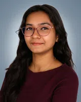 Young woman with long dark hair wearing red glasses and a maroon top smiles confidently against a gray background.