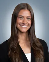 Smiling young woman with long brown hair wearing a black blazer and a cross necklace against a gray background.