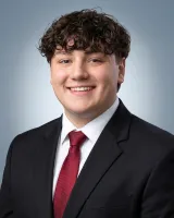 Young man with curly hair smiling in professional black suit, white shirt, and red tie against a light background
