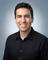 Smiling young man with dark hair wearing a black button-up shirt against a light gray background