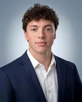 Young man with curly brown hair in navy blazer and white shirt posing against soft blue background.