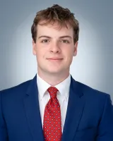 Young man in a blue suit and red patterned tie smiling with a neutral gray background.