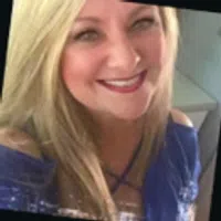 Smiling woman with long blonde hair wearing a blue off-shoulder top indoors.