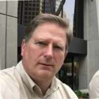 A man in a beige shirt poses outside a modern building with tall windows in an urban setting.