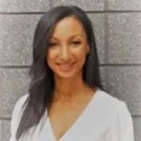 Smiling woman with long dark hair, wearing a white blouse against a gray brick background.