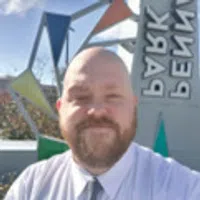 Bearded man in white shirt and tie smiling outdoors with colorful geometric sculpture and sign in background